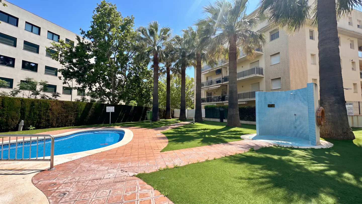 Outdoor pool area with blue water, red brick path, green grass, palm trees, and a blue tiled shower area near a multi-story building.