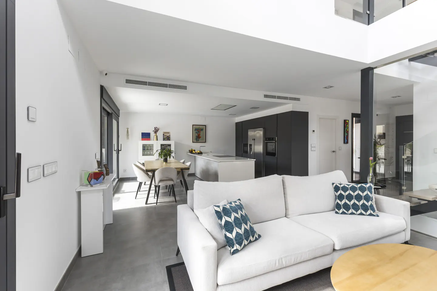 Bright, modern open-plan living space with white sofa, blue pillows, wood table, and gray kitchen cabinets.