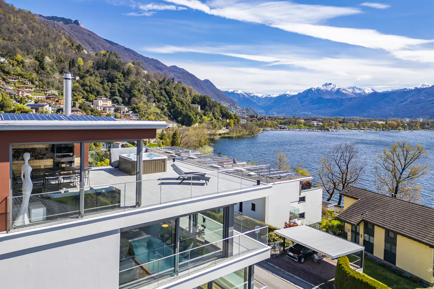 Modern white house with glass balconies overlooks a lake and mountains under a blue sky with clouds.