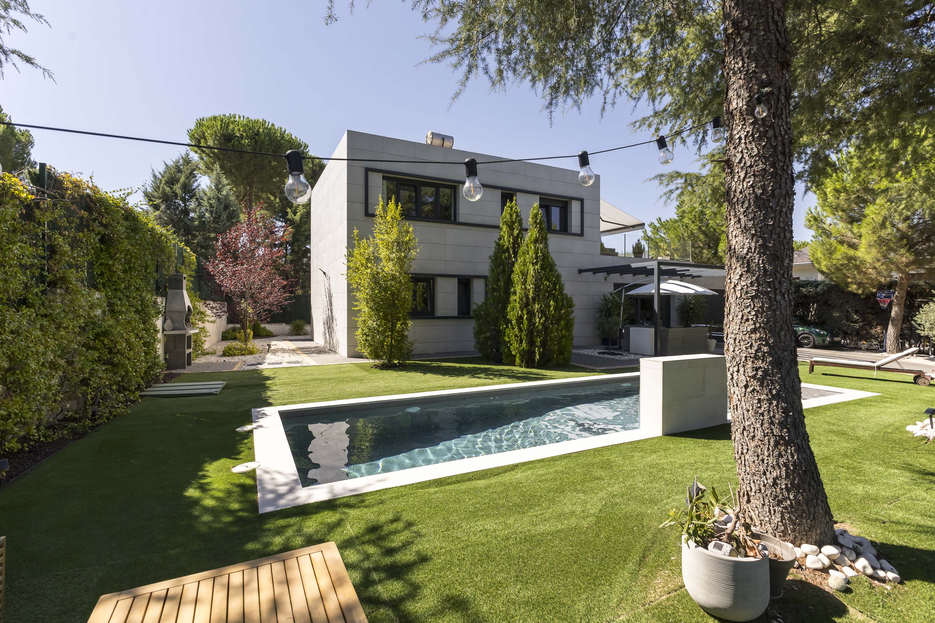 Modern two-story home with a rectangular swimming pool, manicured lawn, and string lights in a sunny backyard. [AI-generated text]