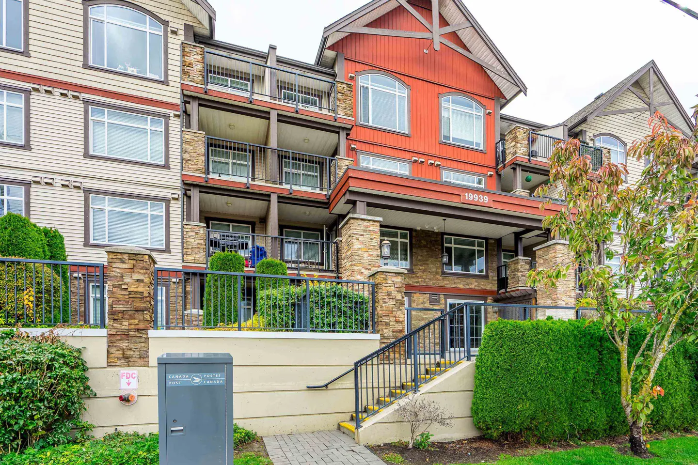 Exterior view of a multi-story condo building with balconies, stone accents, and red siding. A staircase leads to the entrance.