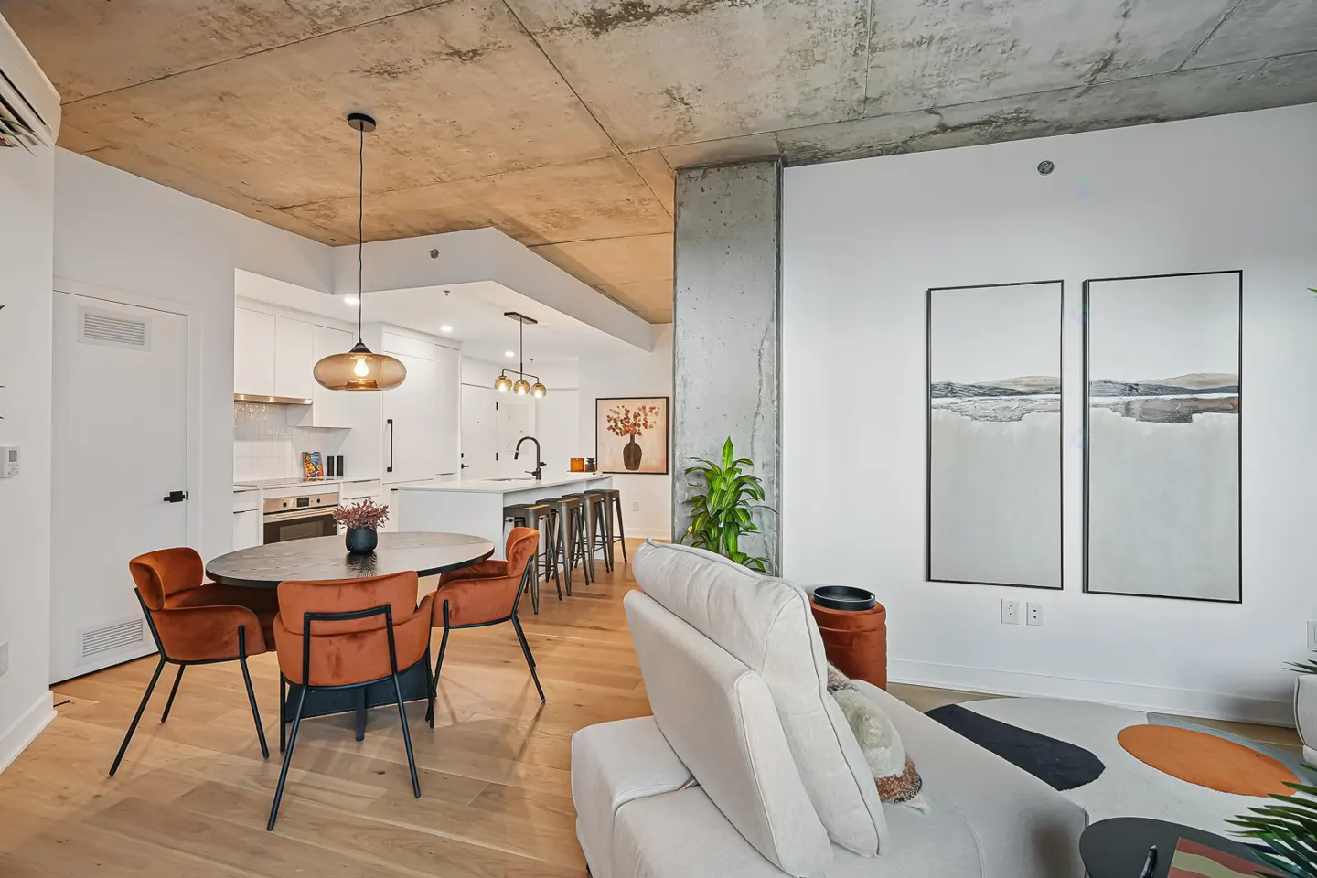 Modern open-concept apartment with concrete ceiling, dining area with orange chairs, and a white kitchen with island.