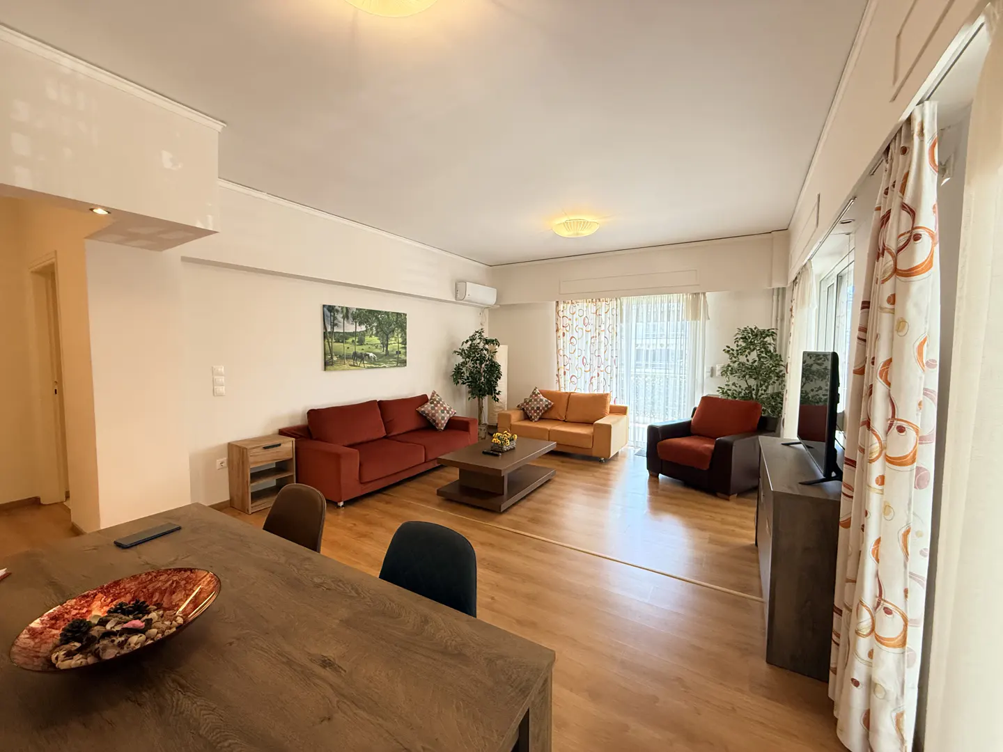 A bright living room with a wood floor, a red sofa, an orange sofa, and a brown armchair. A wood table is in the foreground.