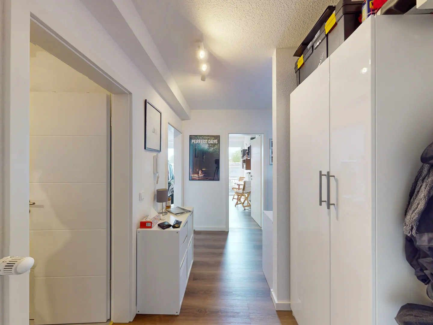 Hallway view with white walls, wood floors, and recessed lighting. A white cabinet and wardrobe are visible. A poster hangs on the wall.