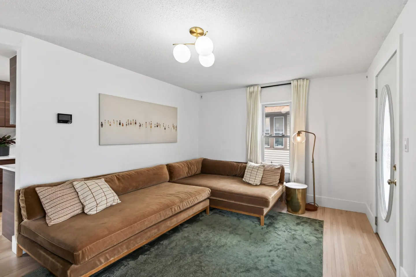 Bright living room with a brown sectional sofa, green rug, white walls, and a modern light fixture. A painting hangs above the sofa.