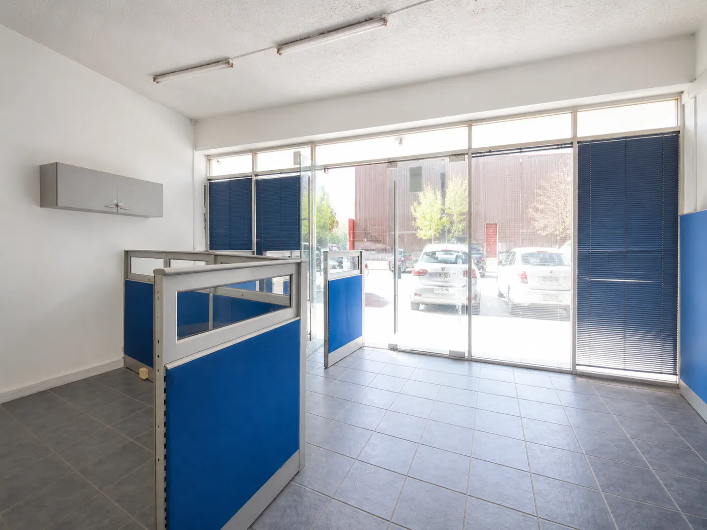 Empty office space with blue cubicle partitions, grey tiled floors, and large glass windows looking out to a parking lot.