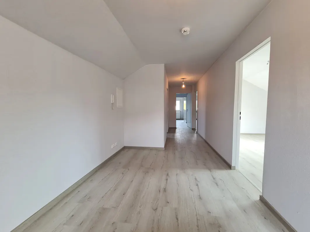 A bright, empty room with light wood floors and white walls. A hallway leads to other rooms. A smoke detector is on the ceiling.