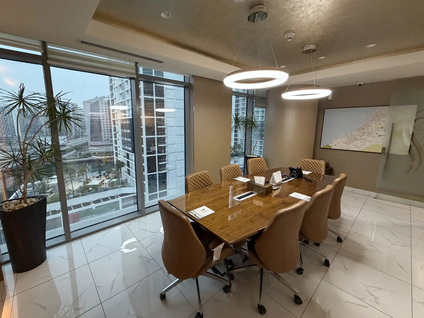 A modern conference room with a wood table, tan chairs, and a city view from a large window. Round lights hang from the ceiling.