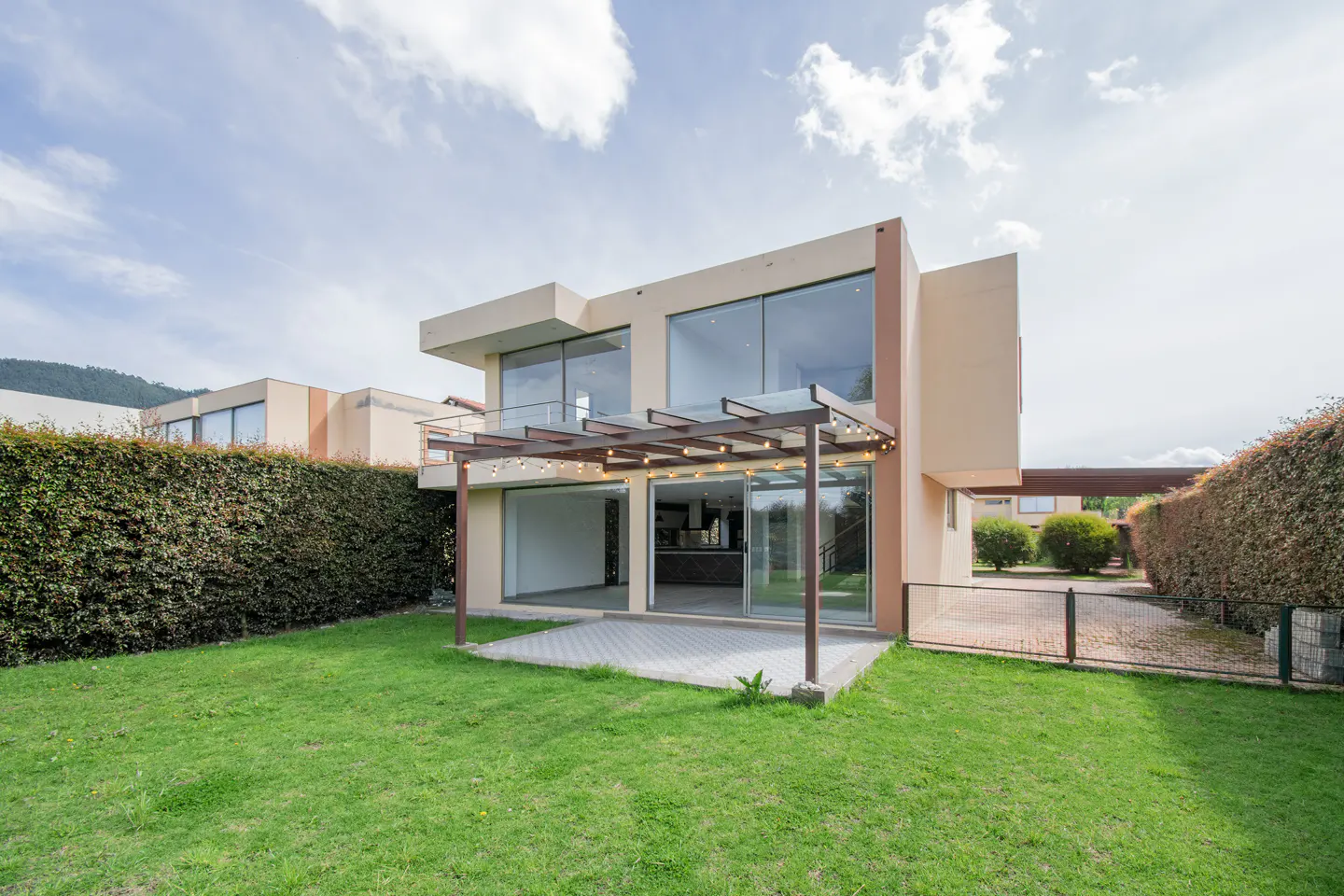 Modern two-story beige house with a backyard, patio, pergola with string lights, and tall green hedges.