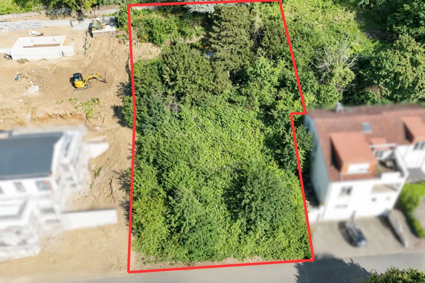 Aerial view of a rectangular plot of land, outlined in red, covered with green trees and bushes. Construction site visible in the background.