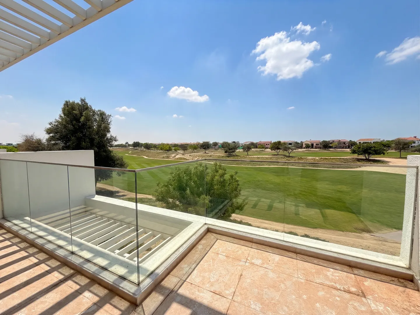 Balcony view of a green golf course under a blue sky. Glass railing and tiled floor. Trees and houses in the distance.