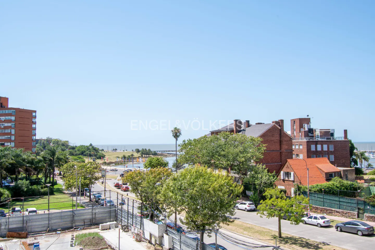 View of a city street with cars, trees, and buildings, leading to a waterfront under a clear blue sky.