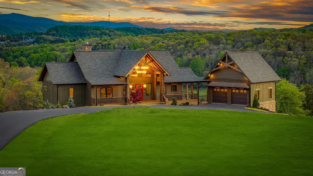 A large, brown, modern house with a gray roof sits on a green lawn with a mountain view at sunset.
