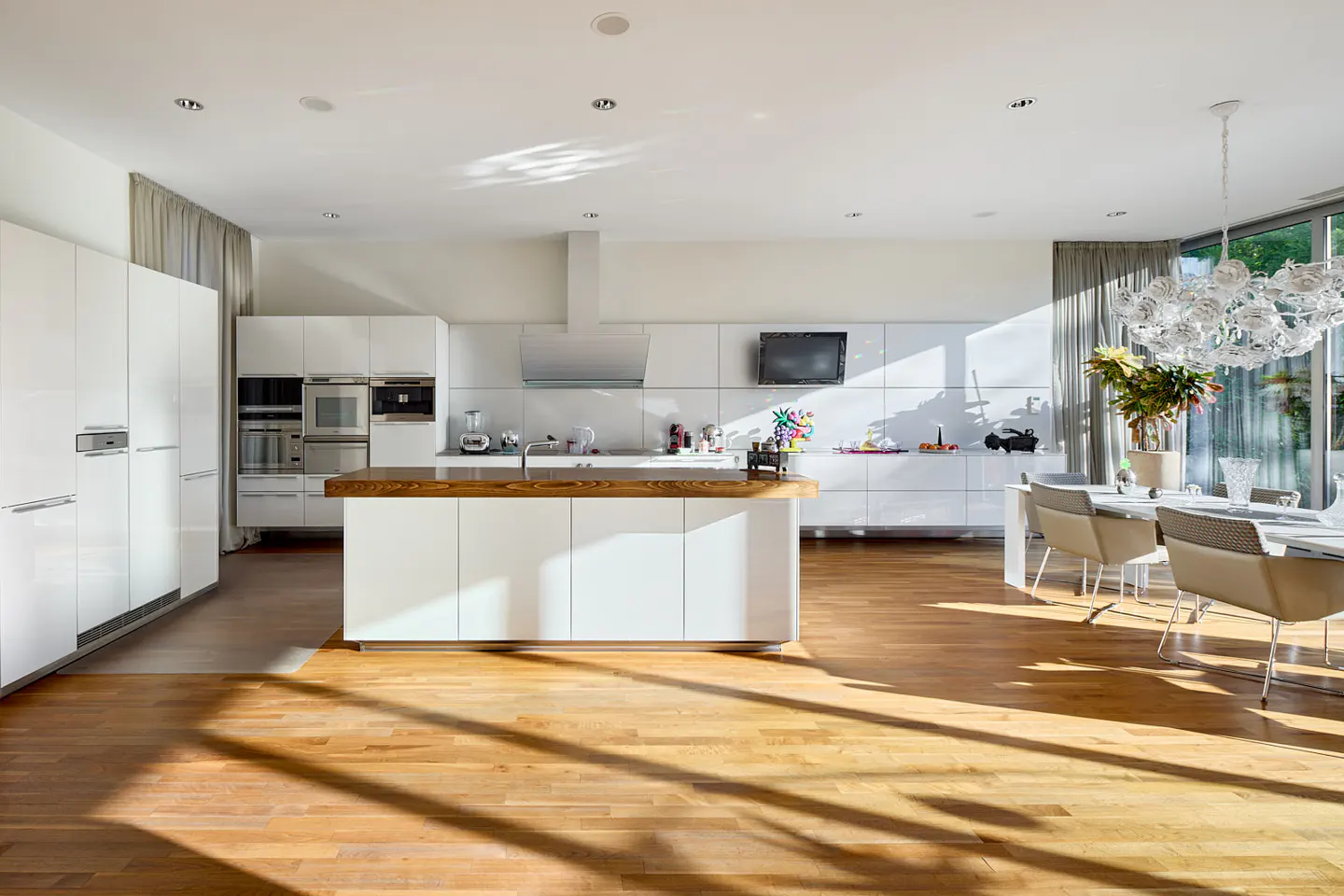 Bright, modern kitchen with white cabinets, wood floors, and a central island. Sunlight streams through the windows onto a dining table.
