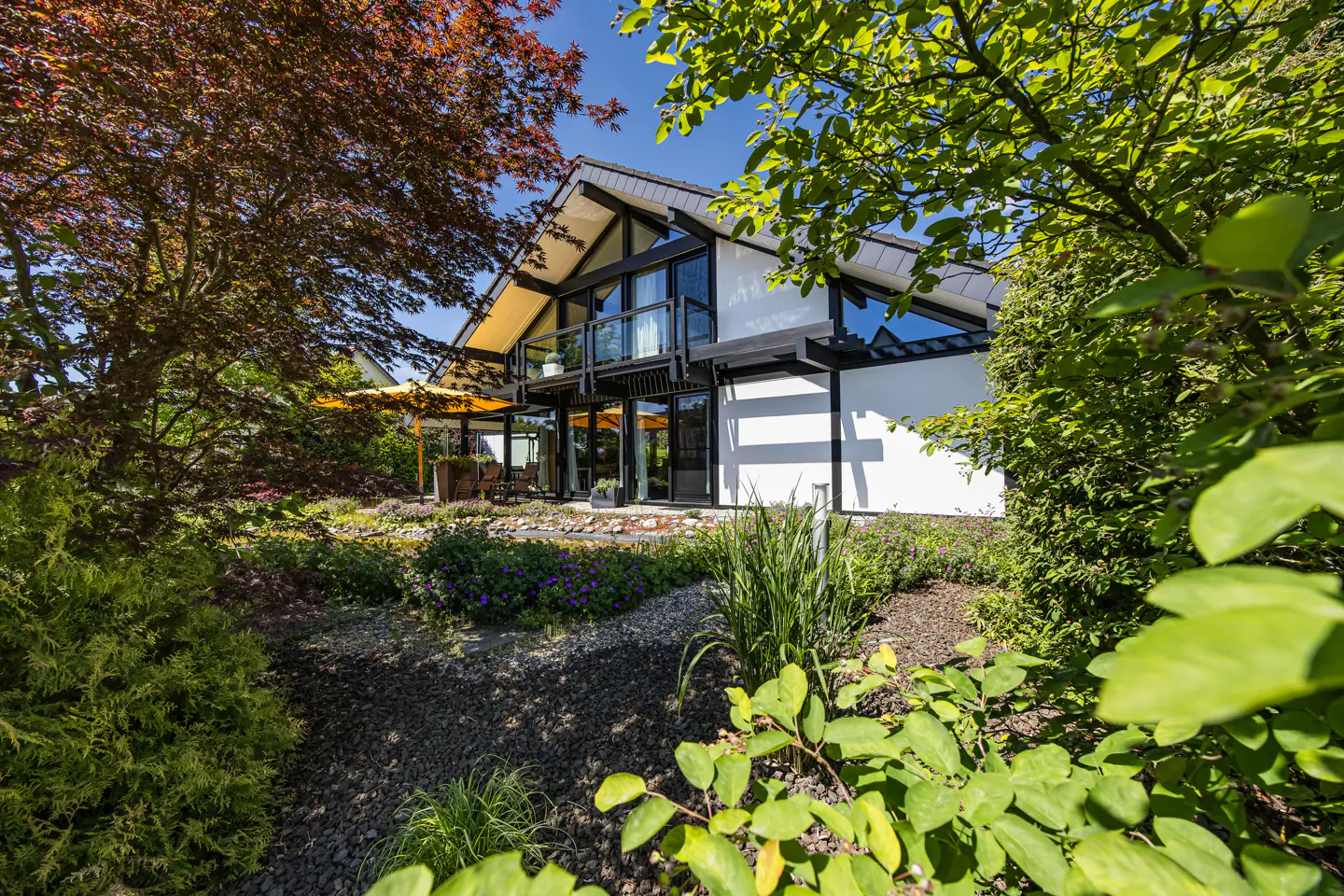 Modern white house with black trim and a balcony, framed by lush green trees and a garden with a yellow patio umbrella.