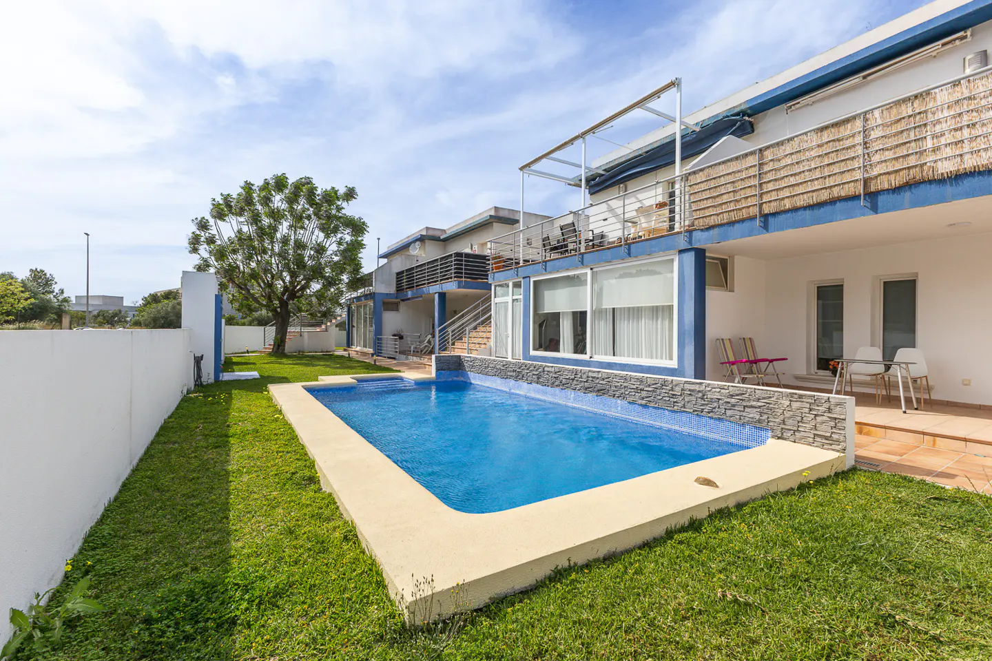 A backyard view of a two-story white house with a blue trim, a pool, and green grass.