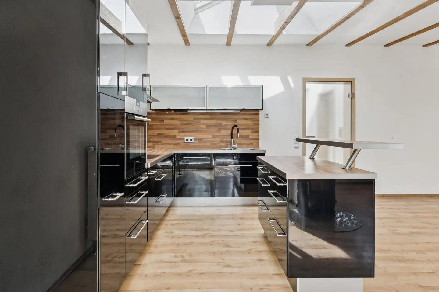 Modern kitchen with black cabinets, wood backsplash, and island. Light wood floors and white walls with exposed beams.
