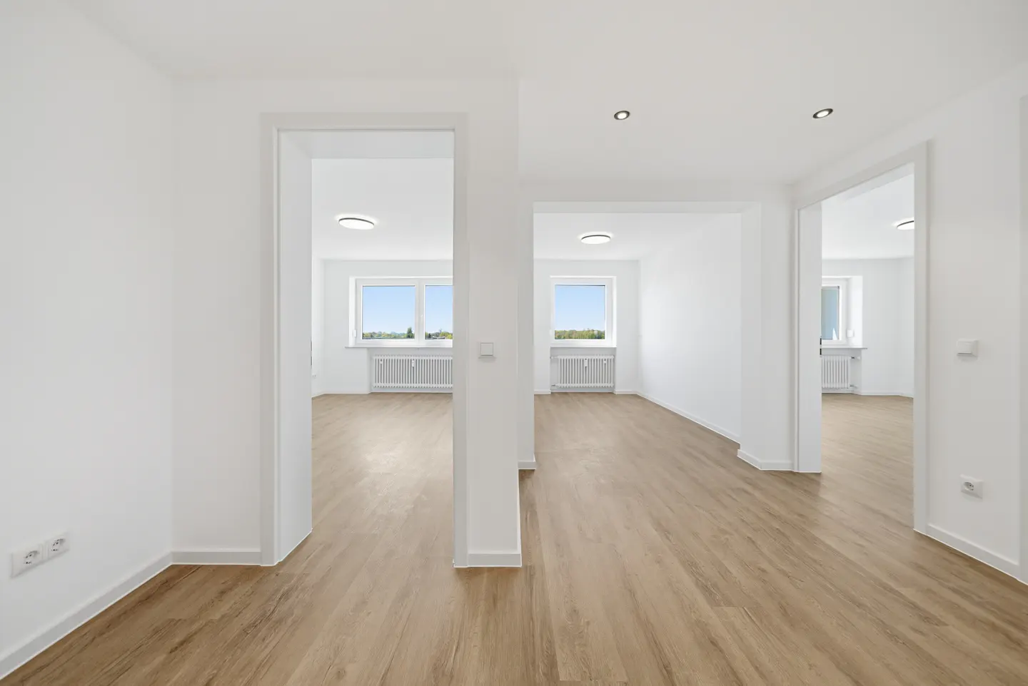 Bright, empty apartment with light wood floors and white walls. Windows in the background let in natural light.