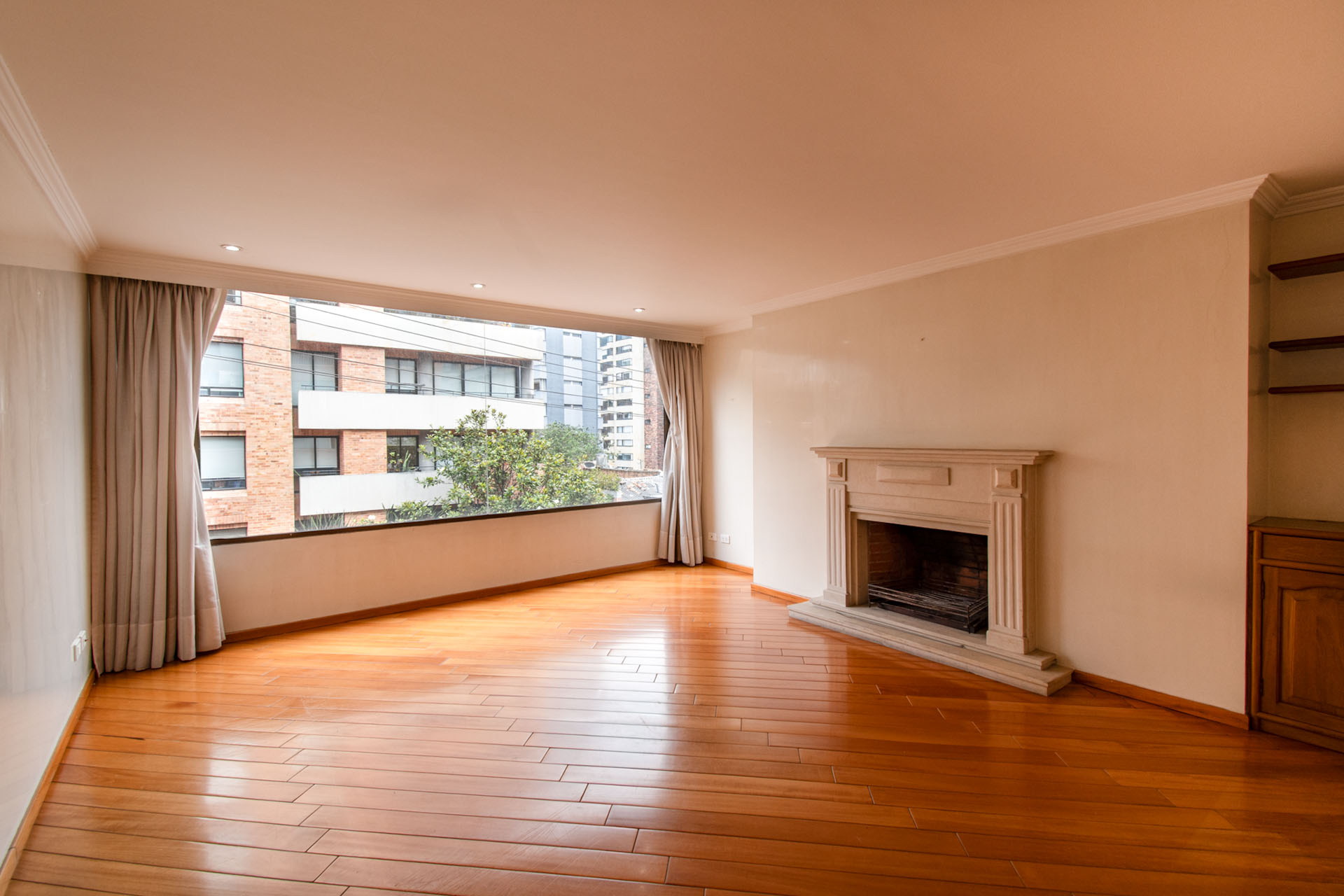 Bright, empty living room with hardwood floors, a stone fireplace, and a large window with curtains. City buildings are visible outside.