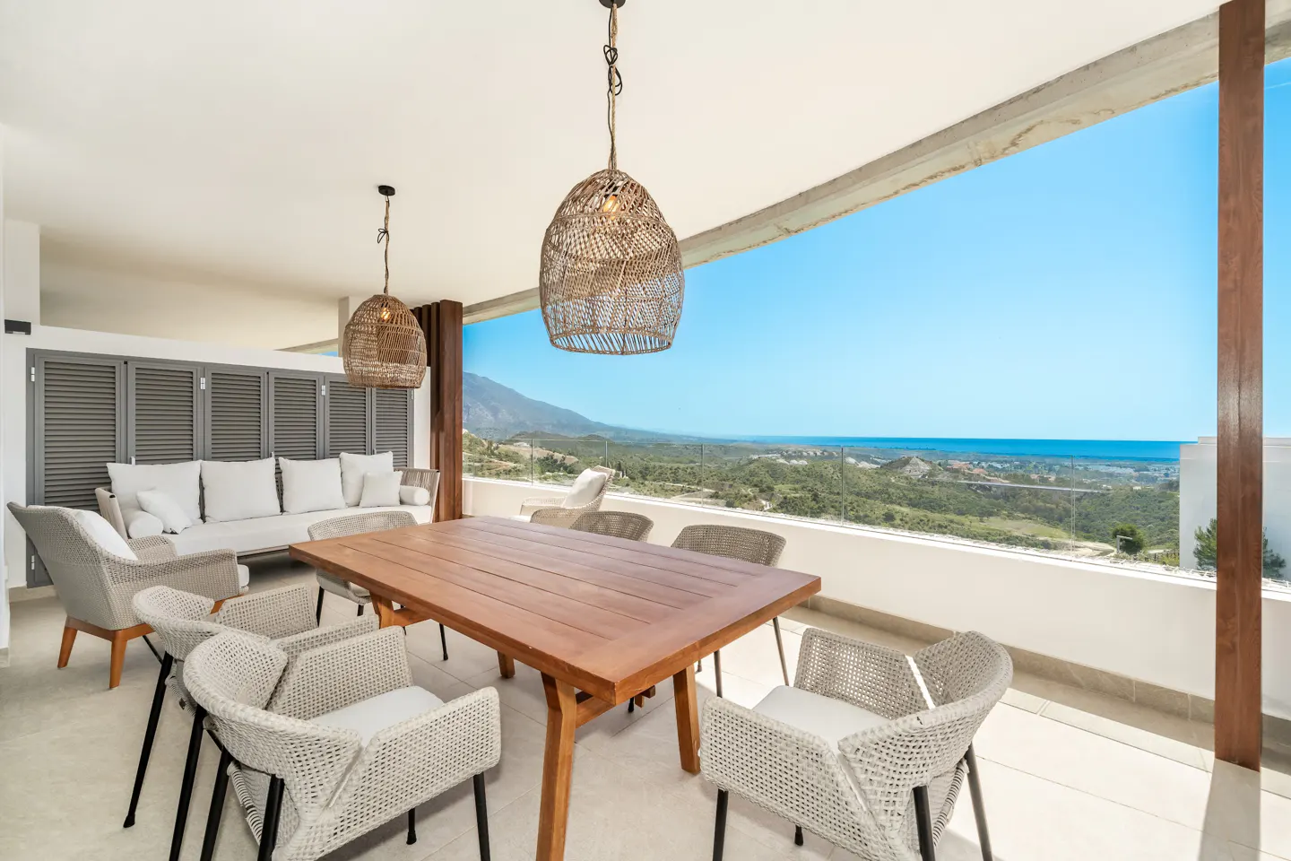 Outdoor patio with a wooden table, wicker chairs, and a white sofa. Wicker pendant lights hang above, with a scenic view of mountains and the ocean.
