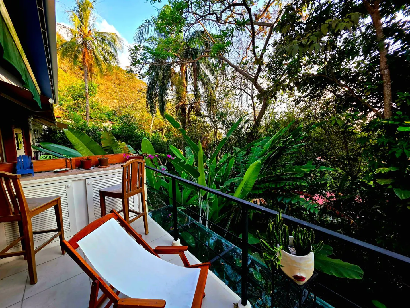 Balcony view with a white lounge chair, wooden chairs, and lush green foliage. A hillside and blue sky are visible in the background.