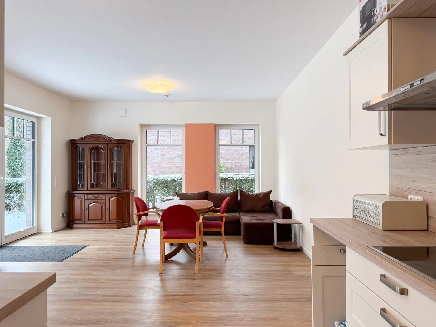 A bright, open-concept living space with wood floors, a brown sofa, a round table with red chairs, and a wooden cabinet.