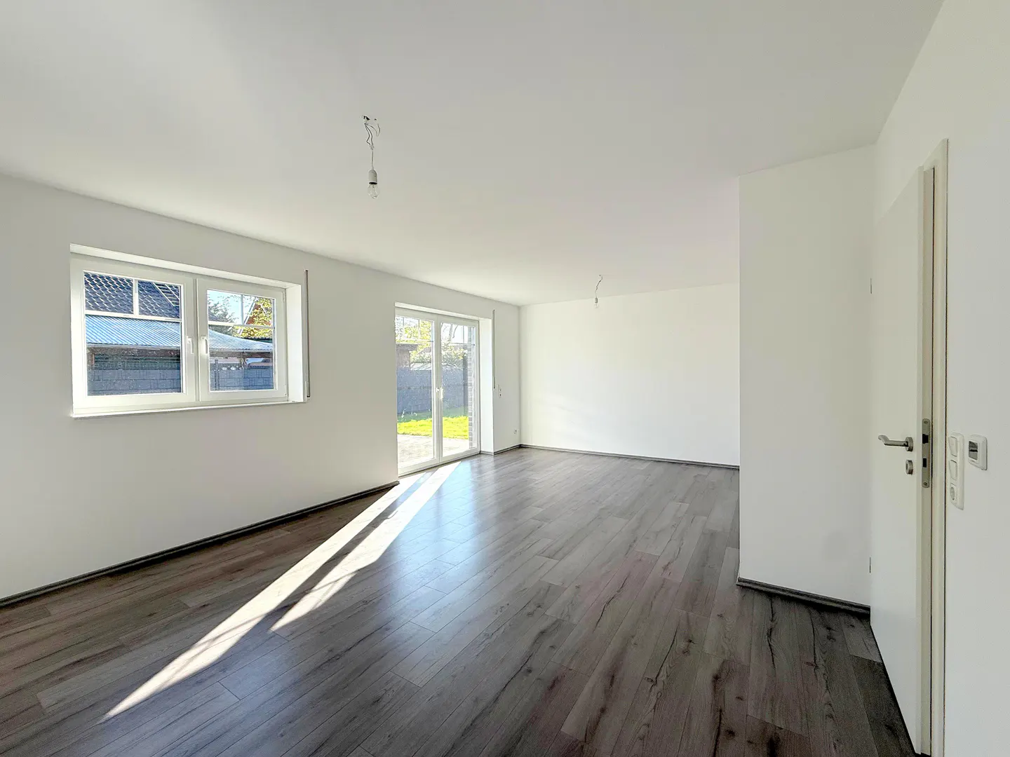 Bright, empty room with dark wood floors, white walls, window, and sliding glass door to the backyard.