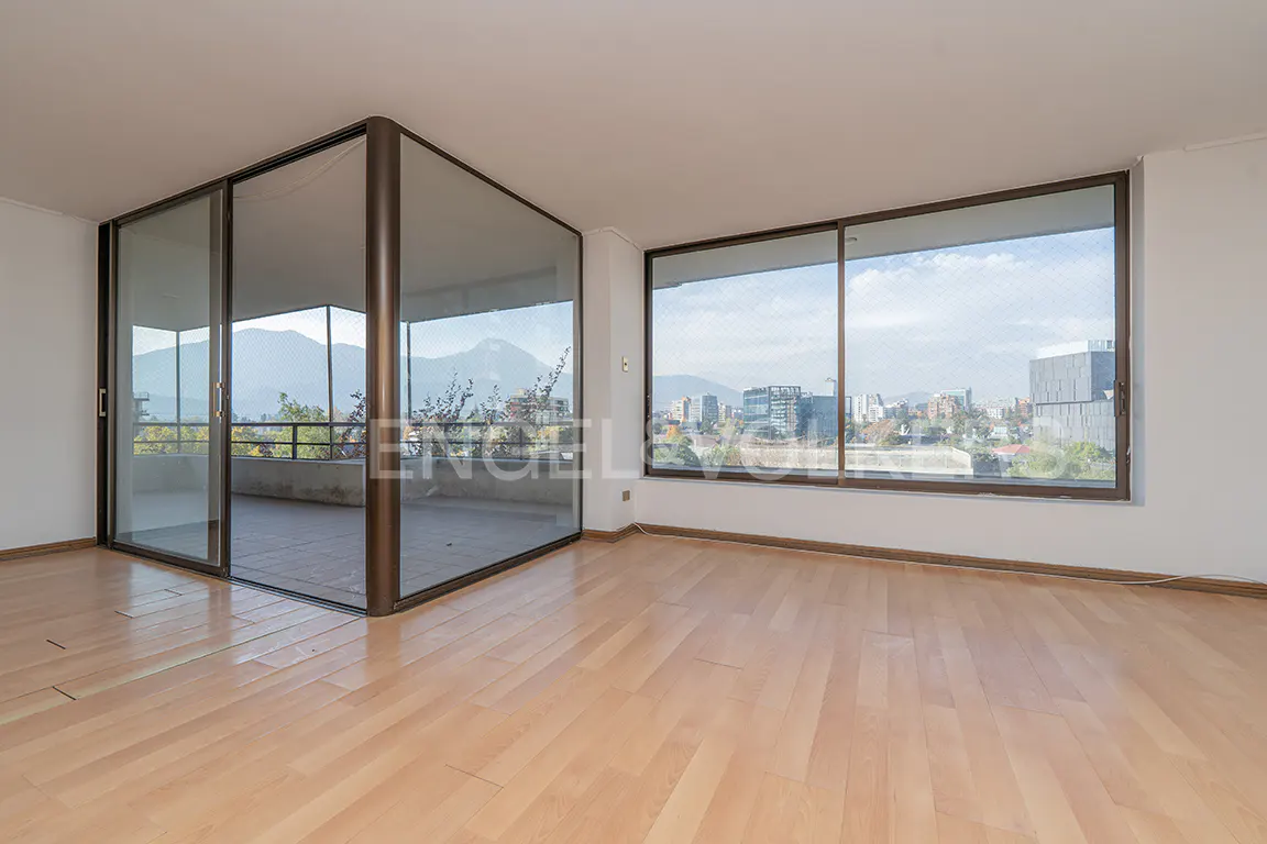 Empty room with light wood floors, white walls, and large windows. Balcony and city views are visible through the windows.