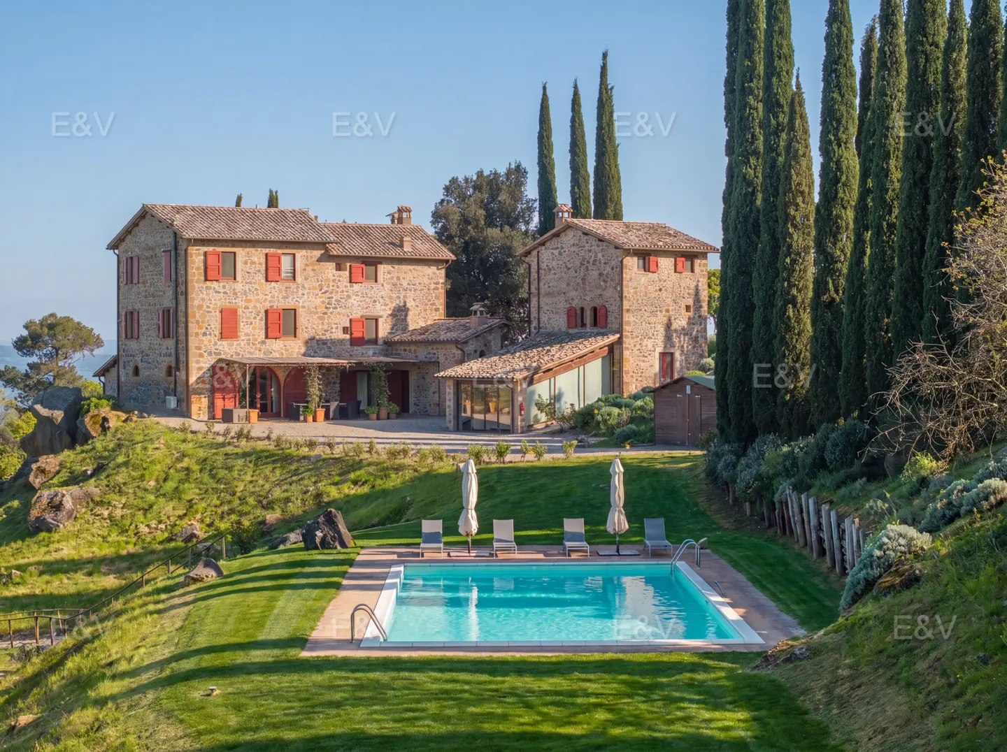 Stone villa with red shutters overlooks a pool with lounge chairs on a green lawn. Tall cypress trees line the property.