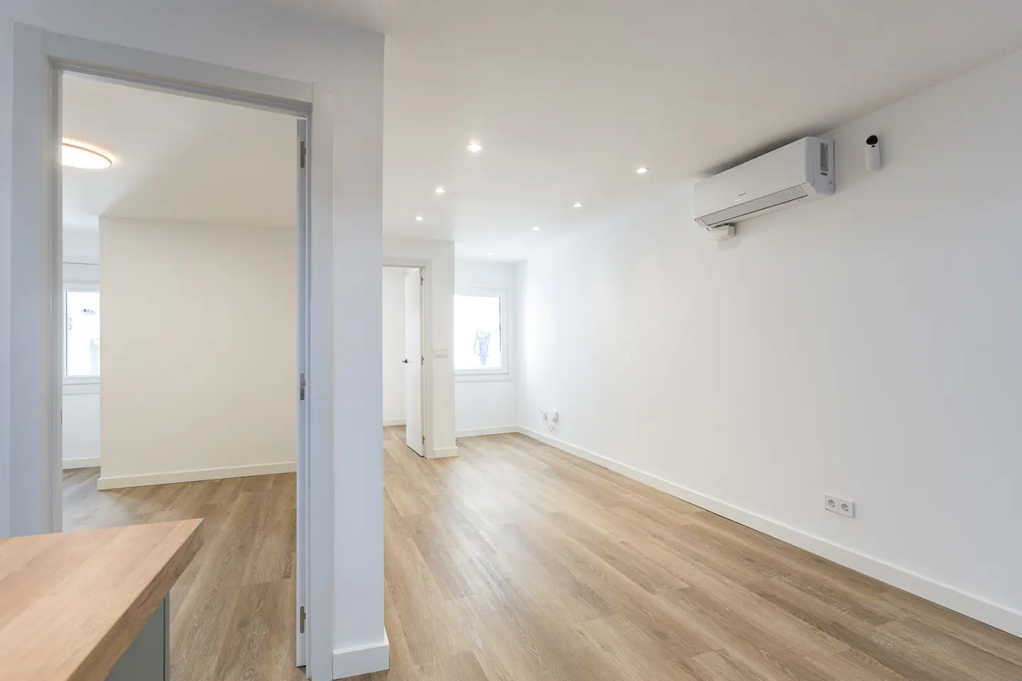 Bright, empty room with light wood floors and white walls. An air conditioner is mounted high on the wall. A doorway leads to another room.