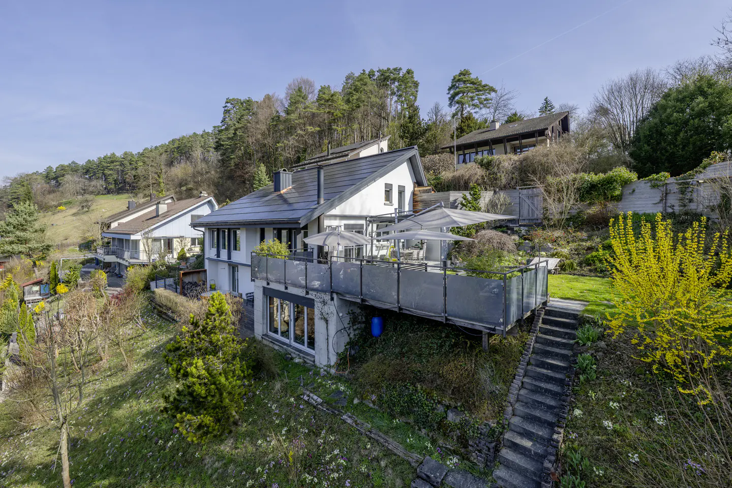 A modern white house with a gray roof sits on a green hillside under a blue sky. A deck with umbrellas extends from the house. Stone steps lead up the hill.