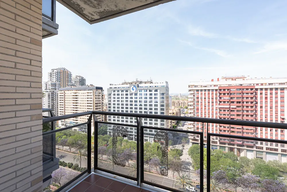 Balcony view of a city with tall buildings, trees, and a street below. The balcony has a brown tile floor and a glass railing with a decorative pattern.