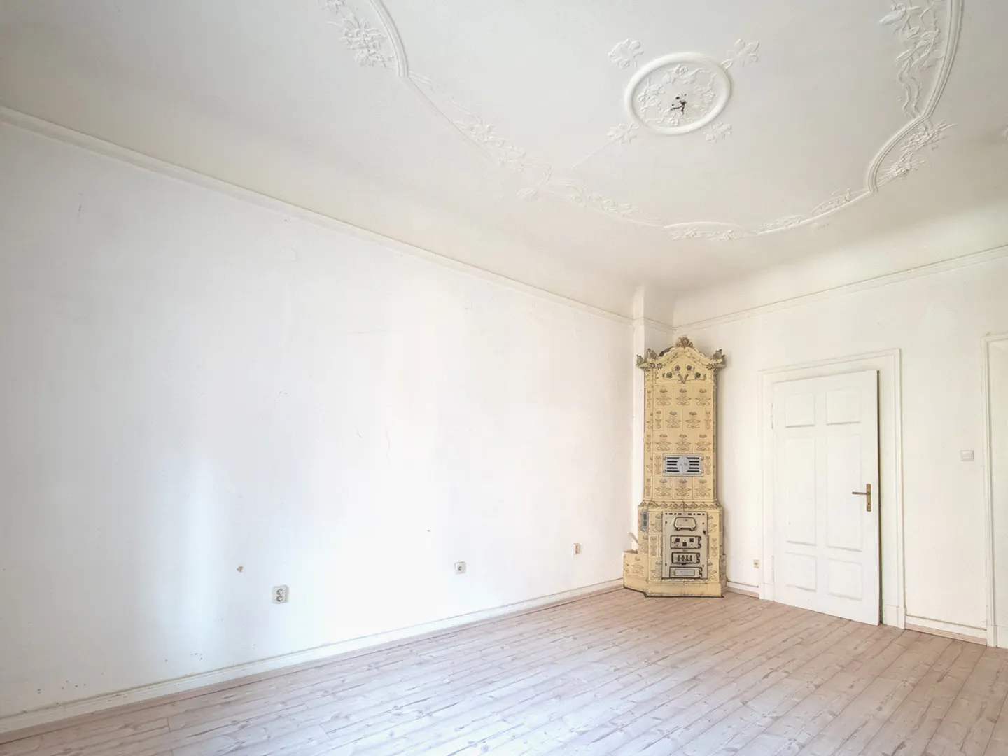 Bright, empty room with white walls, light wood floors, and ornate ceiling. A decorative, tiled stove stands in the corner near a white door.