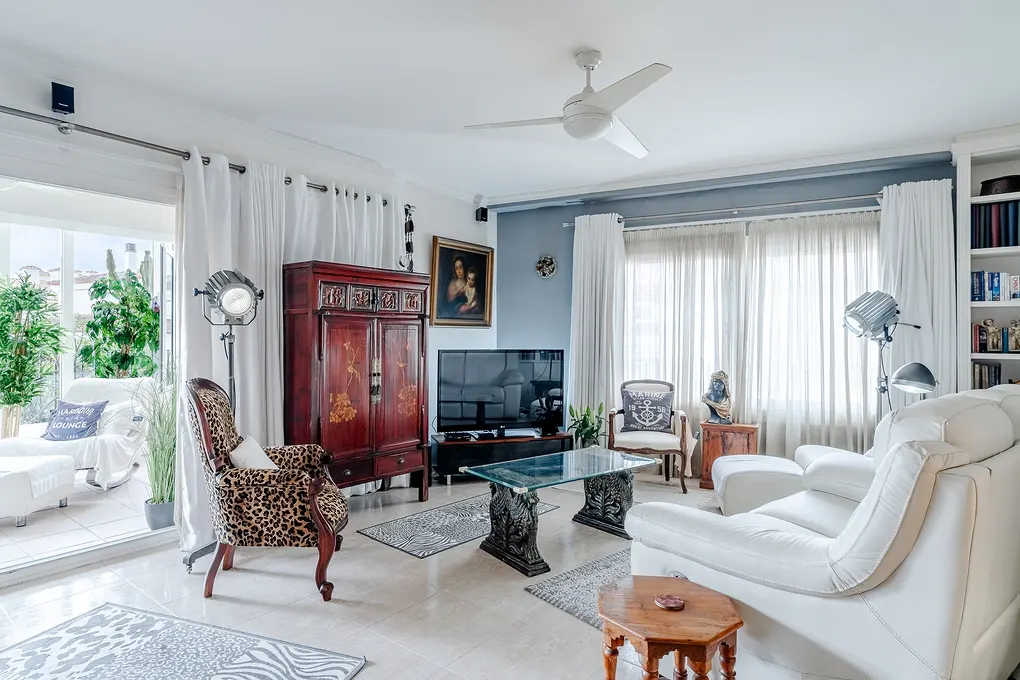 Bright living room with white sofa, leopard print chair, and dark wood cabinet. White curtains frame the windows and open patio.