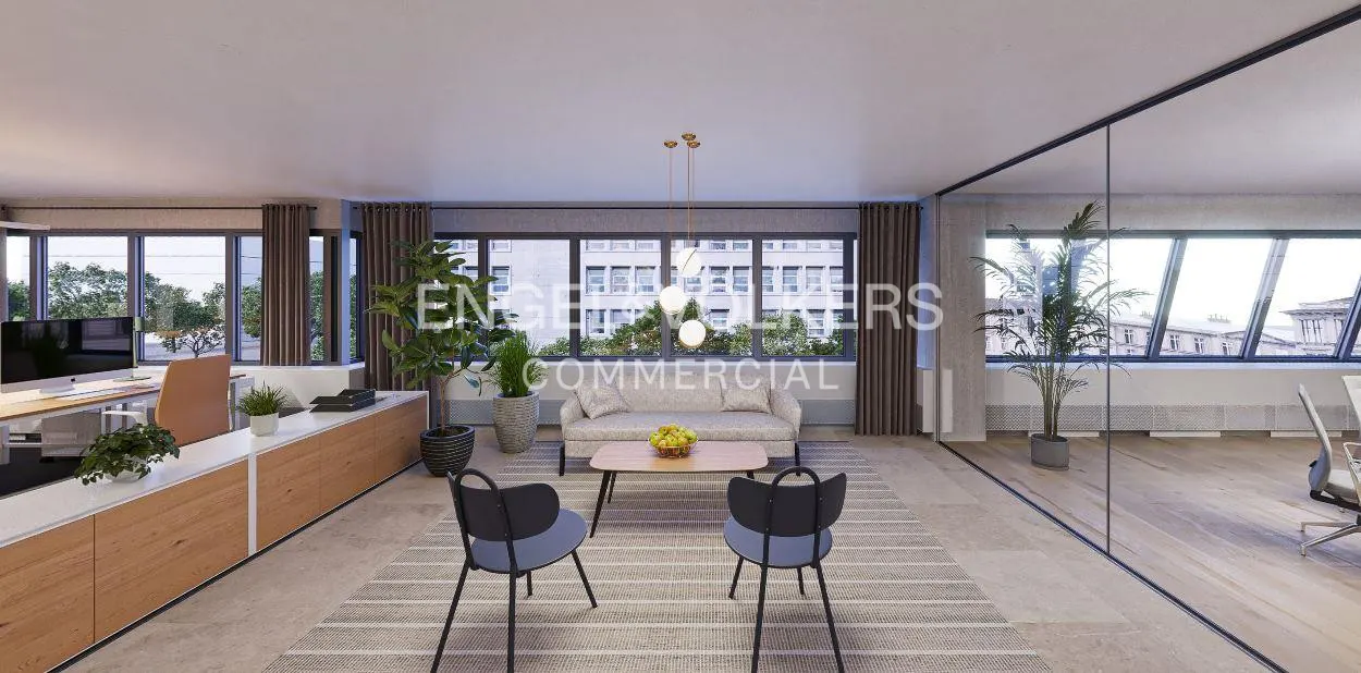Office interior with a sofa, table, and chairs on a striped rug. Large windows offer city views. A desk and plants add to the modern workspace.