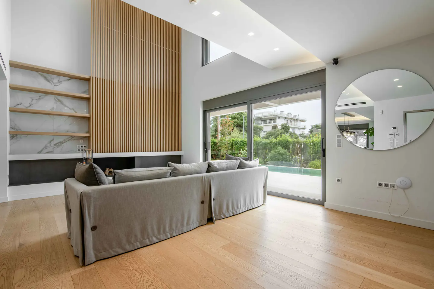 Bright living room with wood floors, a gray sofa, and a wall with wood slats and marble shelves. Sliding glass doors lead to a pool.