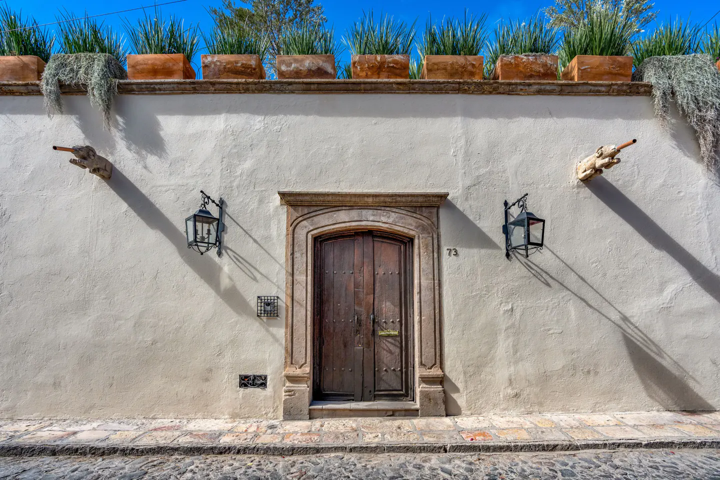 Exterior of a white building with a dark wood door, stone trim, and planters on the roof. Two black lanterns flank the door.