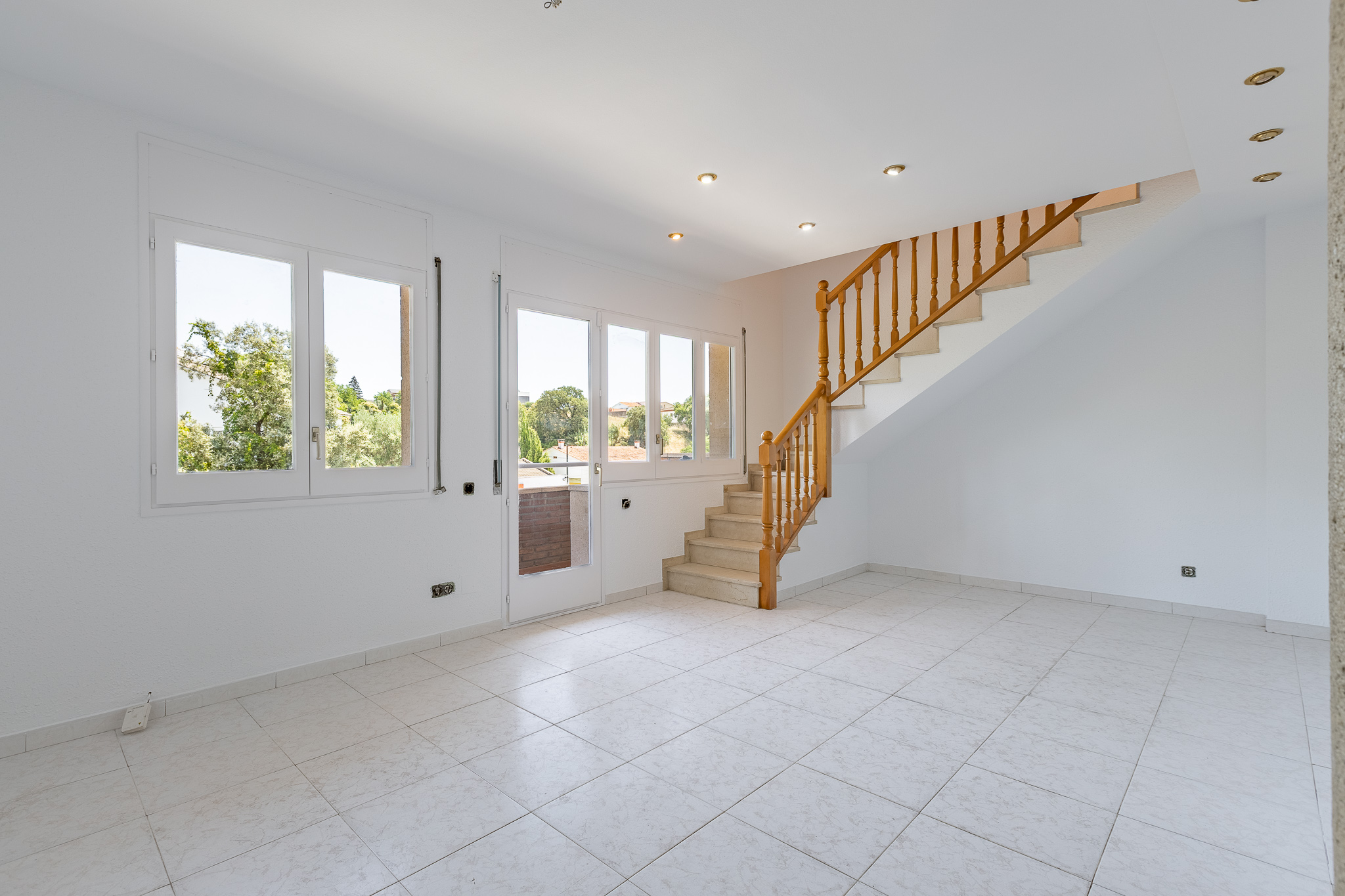 Bright, empty room with white walls, tiled floors, large windows, and a wooden staircase leading to an upper level.