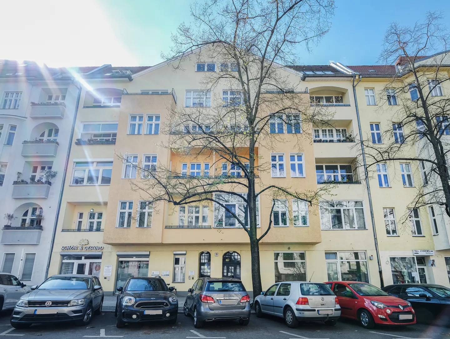Street view of a yellow apartment building with parked cars and a bare tree in front.