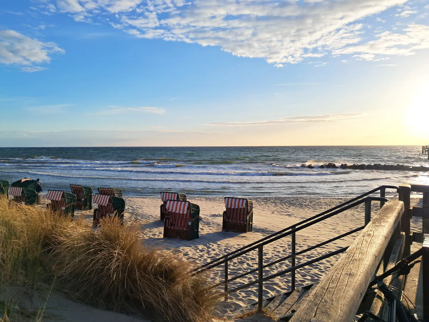 Striped beach chairs on a sandy beach at sunset, viewed from a wooden staircase with dunes in the foreground.