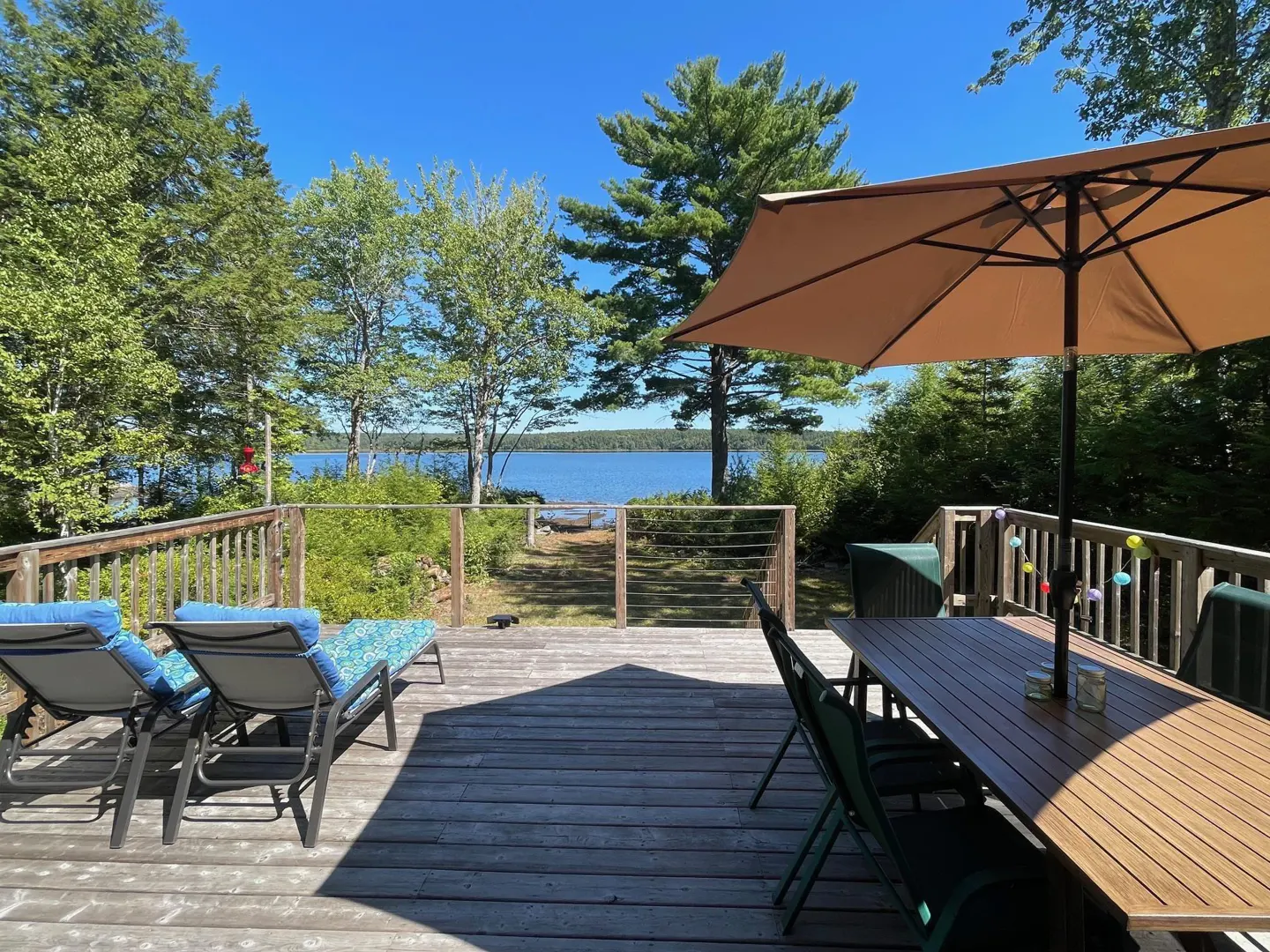Wooden deck with lounge chairs and a dining table under a tan umbrella, overlooking a lake surrounded by lush green trees.