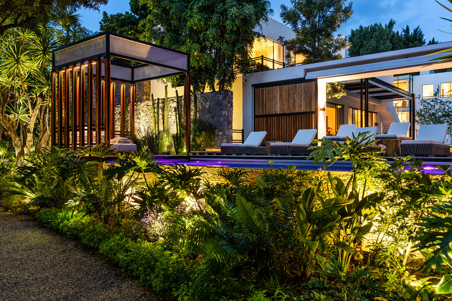 Outdoor view of a modern home with a pool, lounge chairs, and a gazebo at dusk. Lush green plants surround the area, illuminated by warm lighting.