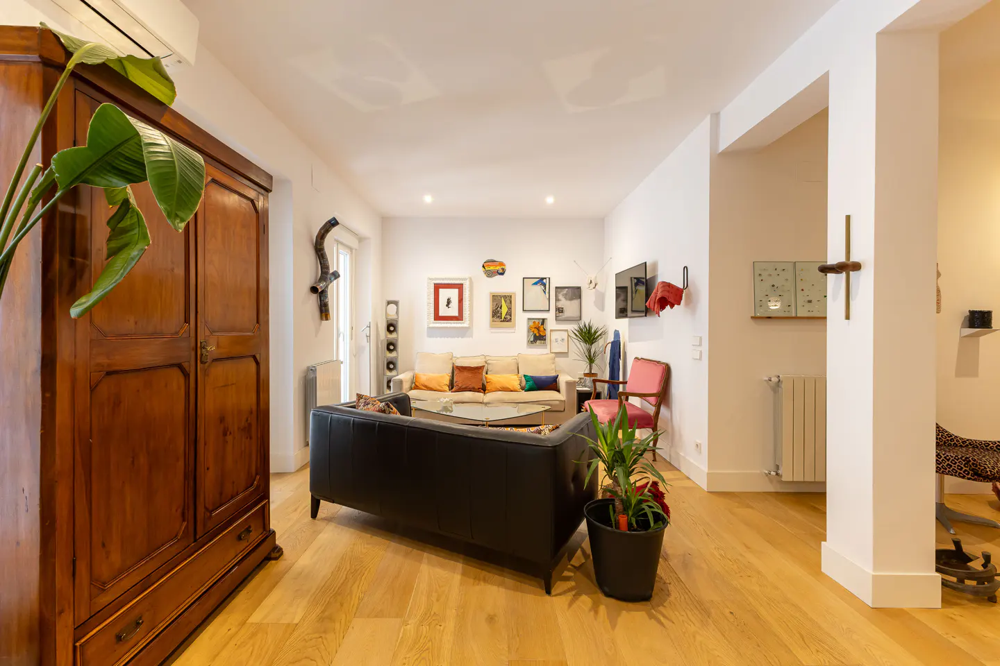 Bright living room with wood floors, white walls, and eclectic art. A large wooden wardrobe stands to the left. Sofas and plants add color.