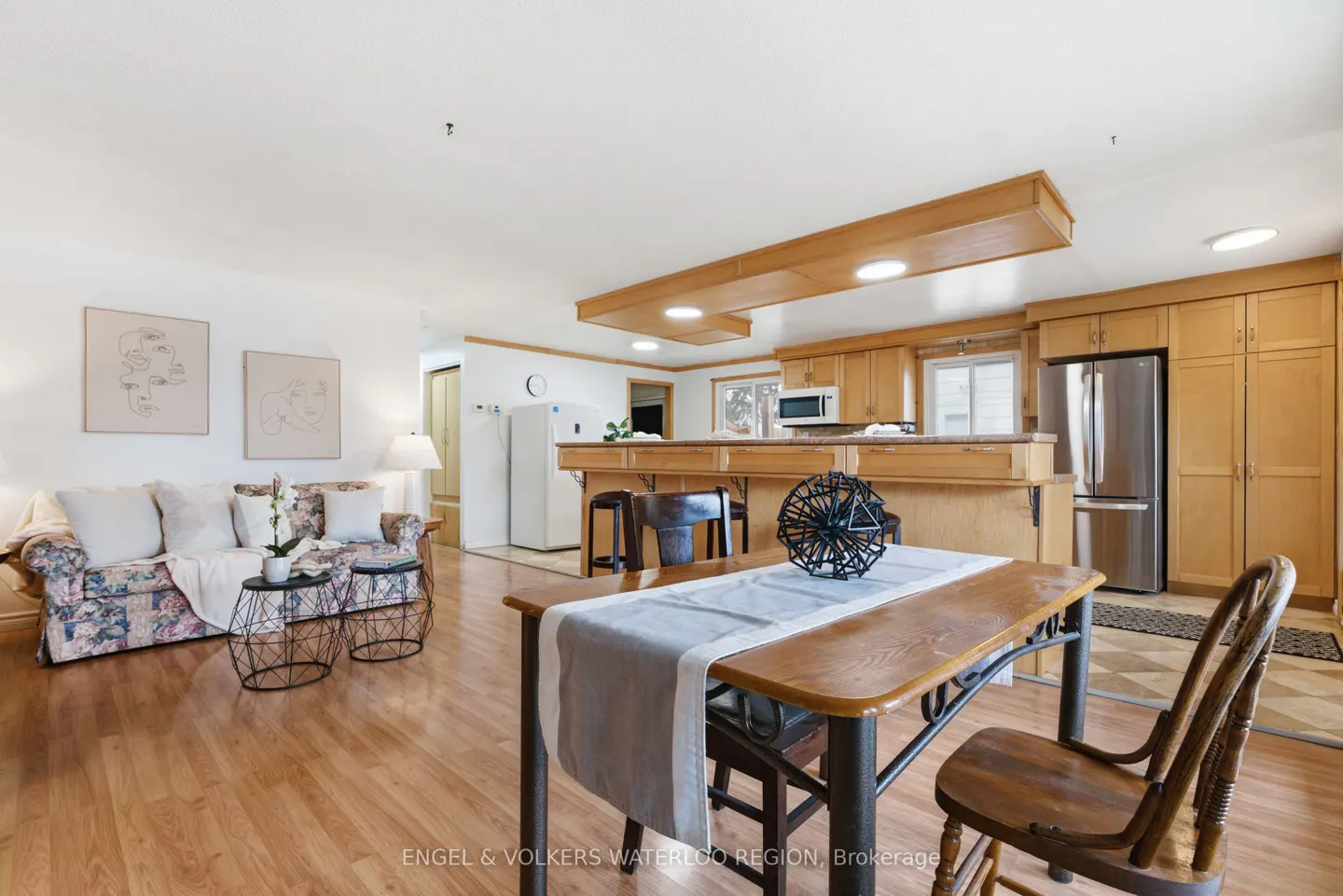 Open-concept living space with wood floors, a floral sofa, and a dining table with a gray runner. The kitchen has wood cabinets and stainless steel appliances.