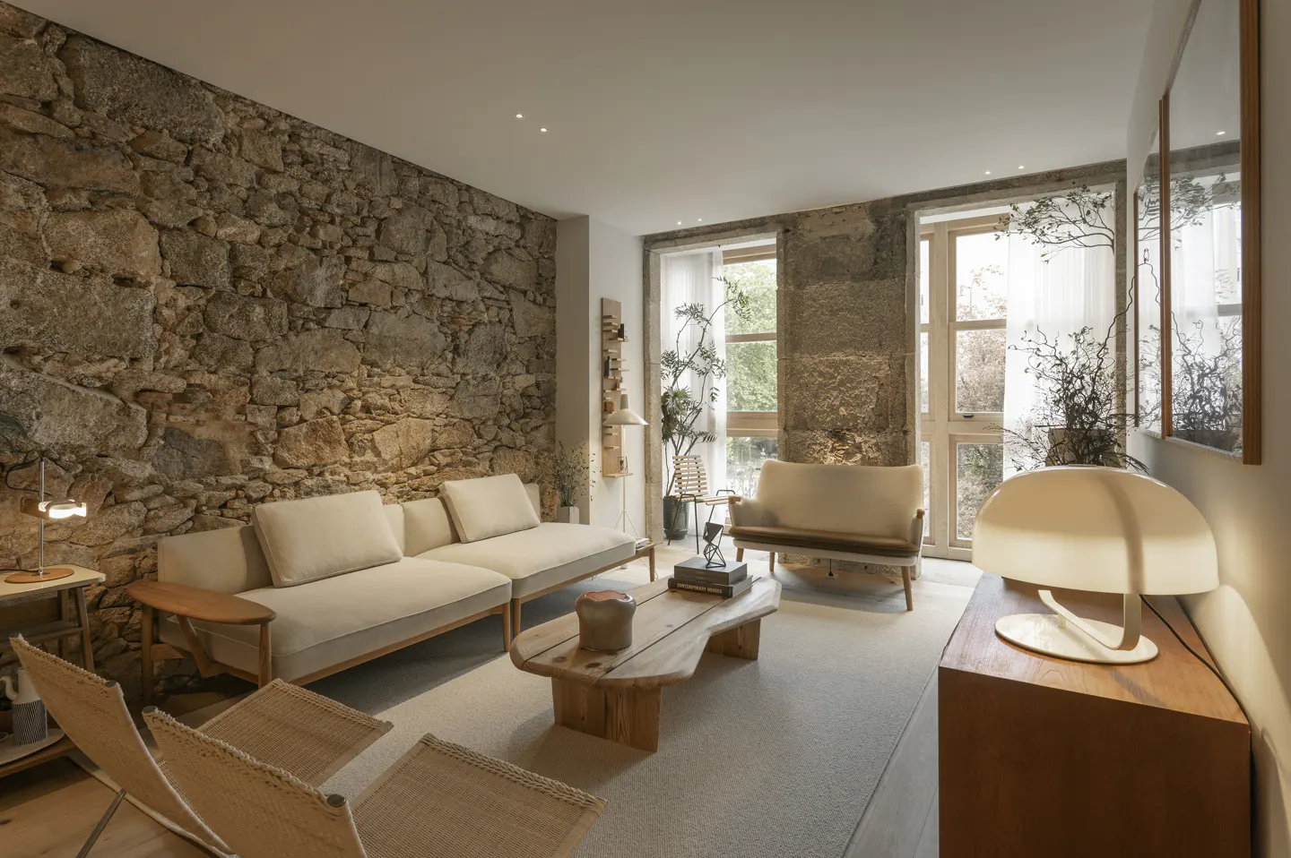 Living room with stone walls, light sofa, wood table, and chairs on a light carpet. Windows with sheer curtains let in natural light.