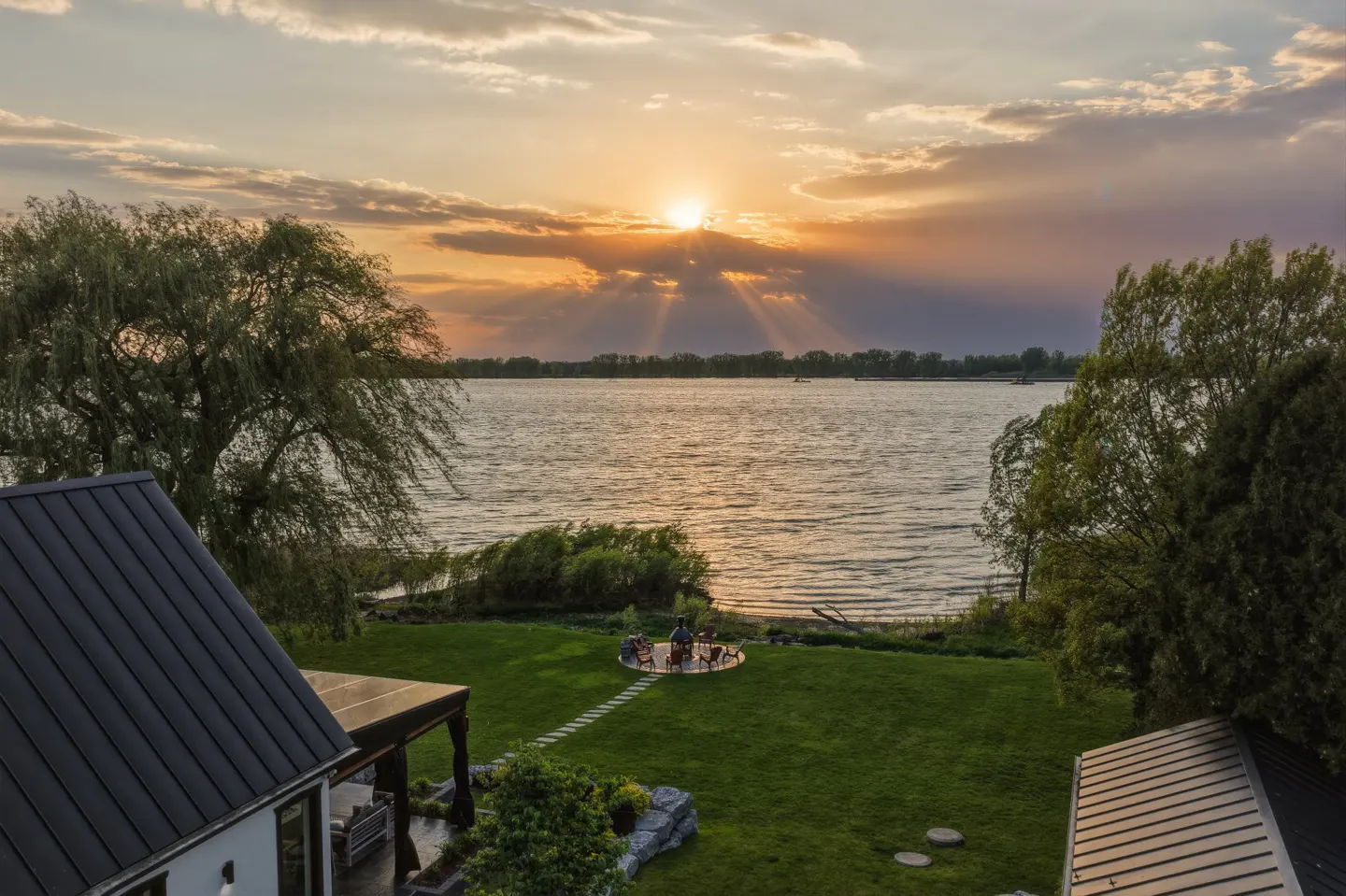 Sunset view of a waterfront property with a green lawn, patio with people, and a house with a dark roof.