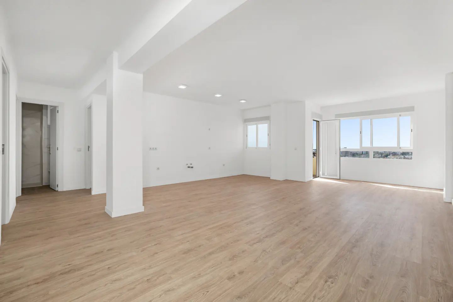 Bright, empty room with light wood floors, white walls, and large windows with a city view.