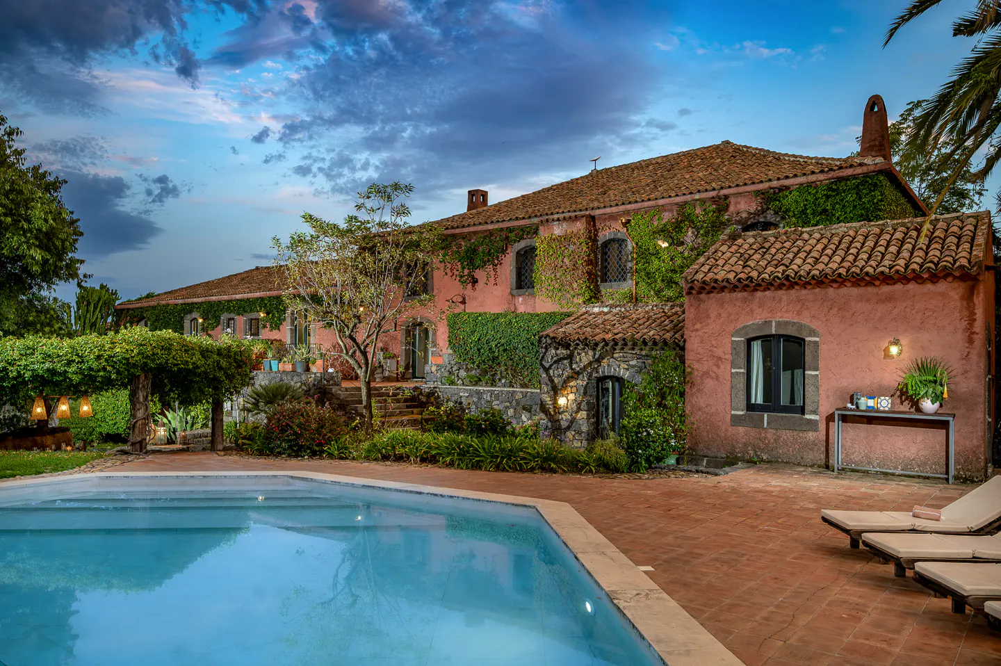 A terracotta villa with ivy-covered walls, a swimming pool, and lounge chairs on a patio under a twilight sky.