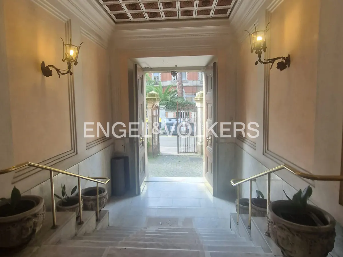 Interior view of a building entrance with marble stairs, brass railings, and potted plants. An open doorway leads to an outdoor view of trees and a gate. Wall sconces provide warm lighting.
