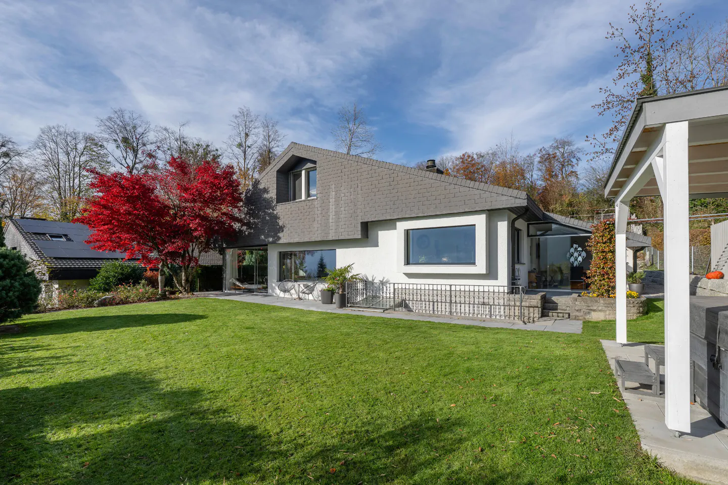 Exterior view of a modern two-story house with a gray roof, white walls, and a large green lawn. A red tree stands out against the blue sky.